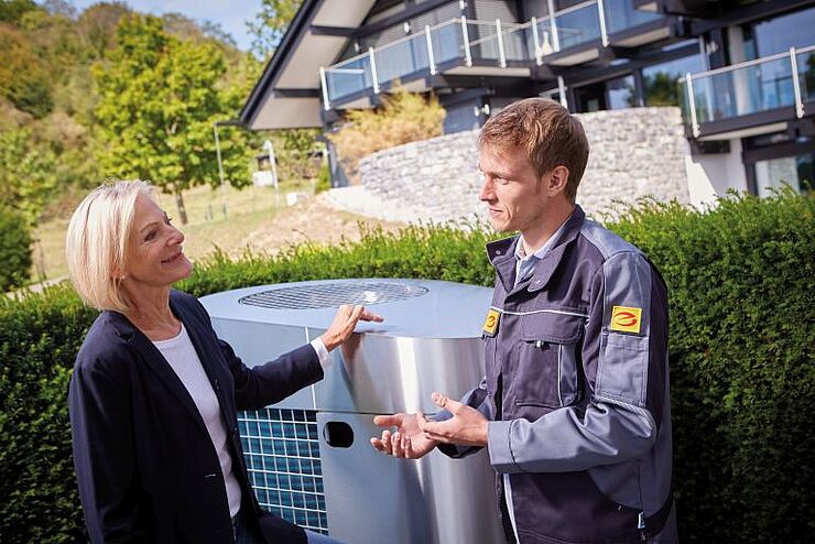 Das Bild zeigt einen Elektrohandwerker mit Kundin vor Wärmepumpe.