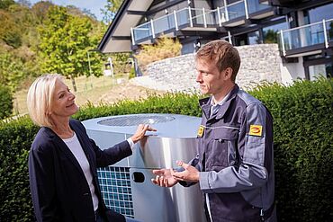 Das Bild zeigt einen Elektrohandwerker mit Kundin vor Wärmepumpe.